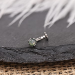 May include: A silver-coloured, flat-backed earring featuring a round, green and white stone. The earring has a post and a small, flat disc at the back. The jewellery is displayed on a dark, textured surface.