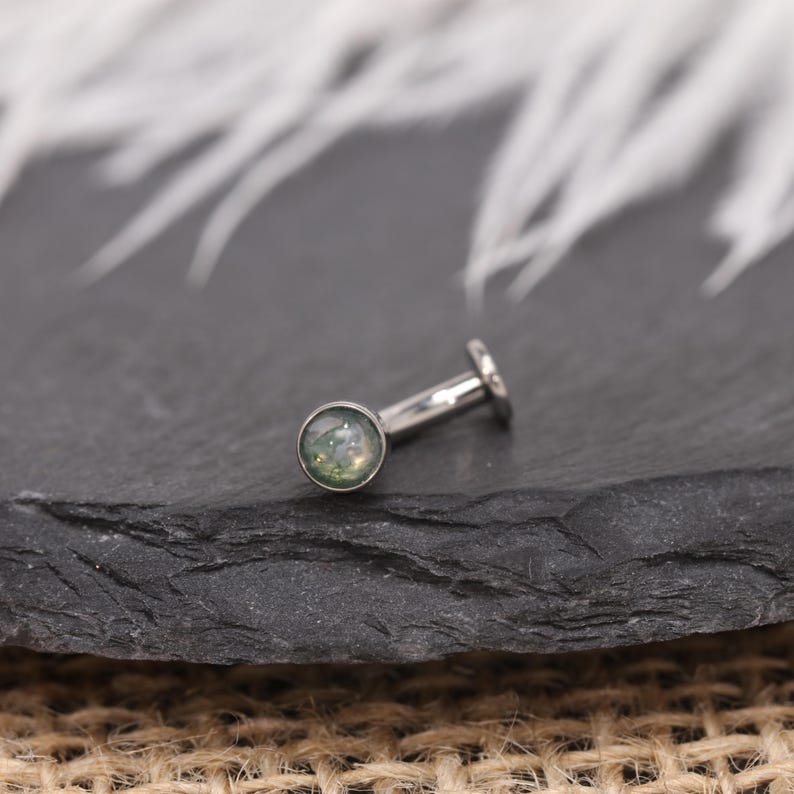 May include: A silver-coloured, flat-backed earring featuring a round, green and white stone. The earring has a post and a small, flat disc at the back. The jewellery is displayed on a dark, textured surface.