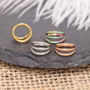 Puede incluir: Anillos de septum en varios colores: oro, plata, iridiscente y oro rosa. Cada anillo presenta un diseño de doble aro. Las joyas se exhiben sobre una superficie oscura y texturizada.