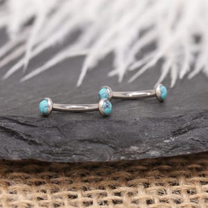 May include: Two curved eyebrow rings with turquoise stone ends. The jewellery is made of silver-coloured metal and is displayed on a dark, textured surface. The background features white, feathery elements.