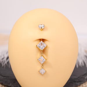 Op de afbeelding: Een goudkleurige navelpiercing met drie bungelende vierkant geslepen heldere edelstenen. Een kleinere, hartvormige heldere edelsteen bevindt zich boven de navel. De sieraden worden getoond op een lichtbruine mannequin.