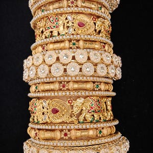 May include: A collection of gold-coloured bangles with detailed patterns. The bangles feature elephant motifs, red and green accents, and small white pearls. The bangles are stacked, creating a layered effect. The bangles are likely made of gold or a gold-toned metal.