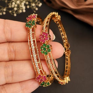 May include: Two gold bangles with floral detailing. The bangles are adorned with red and green gemstones arranged in a flower design. One bangle has a line of small white gemstones. The bangles are set against a dark backdrop.