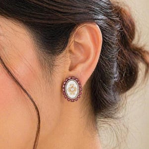 May include: Oval stud earrings featuring a white centre with a gold filigree design, surrounded by small, round, red gemstones. The earrings are set in a gold-coloured metal.