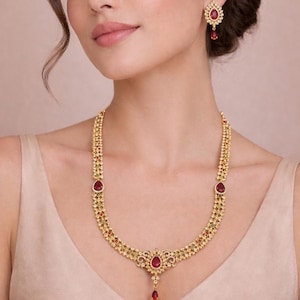 Peut inclure: Un ensemble de collier et de boucles d'oreilles dorées. Le collier présente un pendentif central en forme de goutte d'eau avec une pierre rouge et de petites pierres rouges le long de la chaîne. Les boucles d'oreilles ont un design similaire avec une goutte de pierre rouge.