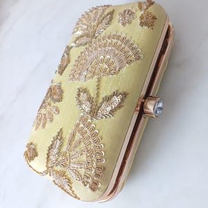 Peut inclure: Une pochette rectangulaire jaune pâle avec des broderies florales dorées et des détails de sequins. La pochette a un cadre en or rose et un fermoir en cristal, un accessoire élégant pour les occasions spéciales.