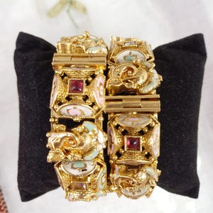 May include: Two gold-tone hinged bangle bracelets with enamel and gemstone accents. The bracelets feature a repeating pattern of ornate squares and floral motifs. The enamel is in shades of pink, green, and white. The gemstones are red.