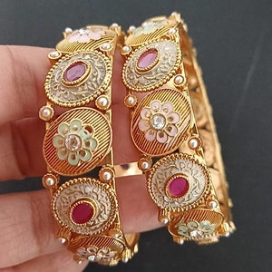Può includere: Due braccialetti d'oro con intricati disegni floreali e accenti di smalto colorati. Ogni braccialetto presenta un motivo ripetitivo di medaglioni rotondi con fiori di smalto rosa, verde e bianco e pietre preziose rosse.