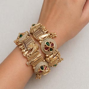 Puede incluir: Brazaletes dorados con intrincados diseños de elefantes y flores. Los brazaletes presentan detalles de gemas verdes y rojas. La joyería se luce en una muñeca, mostrando la detallada artesanía y el estilo ornamentado.