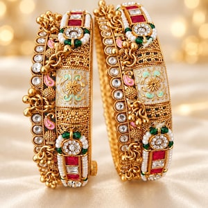 May include: Two gold-toned bangles featuring intricate metalwork, white enamel panels with floral patterns, and embedded red and clear stones. The bangles are accented with small white pearls, green beads, and dangling gold charms along the edges.