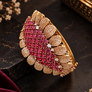 May include: A gold-toned hinged bangle bracelet featuring a semi-circular focal point adorned with oval-shaped deep pink stones and small clear pavé crystals. The metal surface has a textured, granular finish throughout the entire piece.