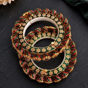 May include: Two ornate gold bangles with red, green, and gold accents. The bangles feature a repeating pattern of red and green diamond shapes and small gold floral details. The inner band is white.