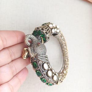 Peut inclure: Un bracelet de style antique orné d'une tête d'éléphant. Le bracelet est de couleur argentée avec des détails complexes, orné d'émeraudes vertes et de pierres précieuses transparentes. La tête de l'éléphant a un œil en pierre rouge et des défenses dorées.