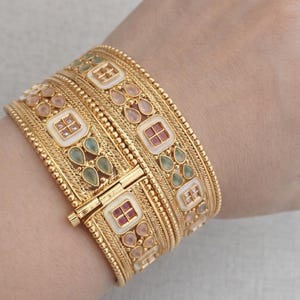 Può includere: Bracciali dorati con dettagli intricati. I bracciali presentano accenti quadrati e a forma di goccia in bianco, rosa, rosso e verde. Il design include una chiusura a fermaglio. I gioielli sono indossati al polso.