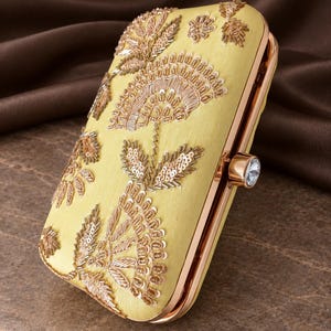 May include: A rectangular pale yellow clutch bag featuring intricate gold-toned beadwork and sequin embroidery in a floral pattern. The bag has a metallic rose gold frame and a single clear crystal stone set into the side clasp mechanism.