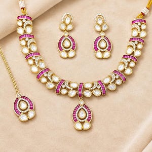 May include: A gold-toned jewellery set consisting of a necklace, a pair of drop earrings, and a maang tikka. Each piece features teardrop-shaped white stones surrounded by small clear crystals and accented with rectangular magenta-coloured stones.