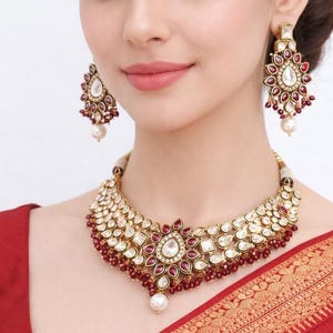 May include: A gold-toned necklace and matching earrings set with clear and red gemstones and a pearl drop. The jewellery is worn with a red garment. The earrings and necklace have a floral design.