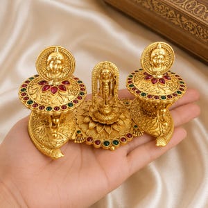 Puede incluir: Conjunto de artefactos religiosos dorados ornamentados. El conjunto incluye tres piezas: dos más grandes, circulares, con un diseño de rostro y una figura central. Cada pieza está adornada con piedras preciosas rojas y verdes. Las piezas se sostienen en una mano.