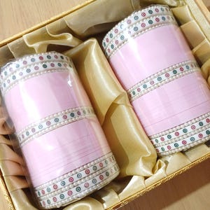 Può includere: Due braccialetti rosa con dettagli decorativi bianchi, verdi e rossi in una scatola dorata con fodera in raso.