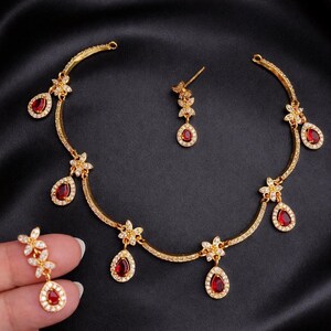 May include: A gold-coloured jewellery set comprising a necklace and earrings. The necklace and earrings feature a floral design with red, teardrop-shaped gemstones and clear accent stones. The jewellery is displayed against a black background.