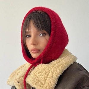 May include: A woman wearing a red knit hooded scarf with a long string tie. She is wearing a brown jacket with a cream colored fleece lining.