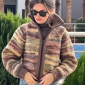 May include: A multi-colored, striped crochet jacket with a zipper closure. The jacket features shades of brown, green, and yellow. The person is wearing sunglasses and blue jeans. The jacket has a high collar.