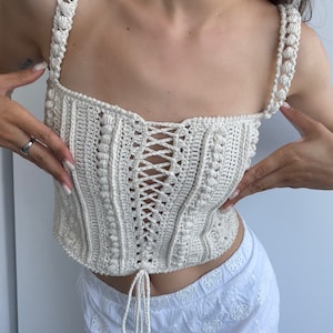 Op de afbeelding: Een wit gehaakt topje met een veter aan de voorkant en een strik in de taille.