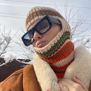 May include: A person wearing a brown leather jacket with a white fur collar, a knitted hat and neck warmer with orange, green, and brown stripes, and black sunglasses.