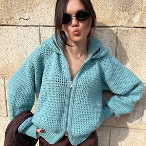 May include: A turquoise, hand-crocheted zip-up hoodie with a hood and long sleeves. The jacket has a silver zipper and is worn over a brown skirt. A brown handbag is held in the left hand. The background is a textured stone wall.