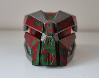 May include: A red and green plastic mask with a silver trim. The mask has a geometric design and is reminiscent of a robot or alien.