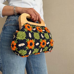 Puede incluir: Bolso de mano de ganchillo con asa de madera. El bolso presenta un estampado floral en naranja, verde y blanco sobre un fondo azul marino. El bolso lo sostiene una persona que lleva una camisa blanca y vaqueros azules.