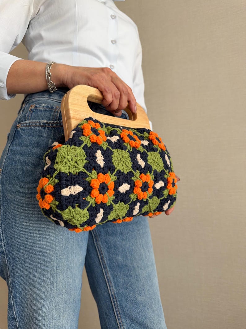 Puede incluir: Bolso de mano de ganchillo con asa de madera. El bolso presenta un estampado floral en naranja, verde y blanco sobre un fondo azul marino. El bolso lo sostiene una persona que lleva una camisa blanca y vaqueros azules.