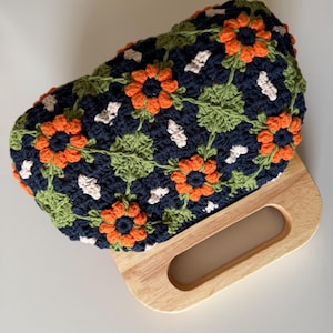 Puede incluir: Un bolso de mano de ganchillo hecho a mano con un estampado floral en azul marino, naranja, verde y blanco. El bolso tiene un asa de madera. El dise&ntilde;o floral incluye flores naranjas, hojas verdes y detalles blancos.