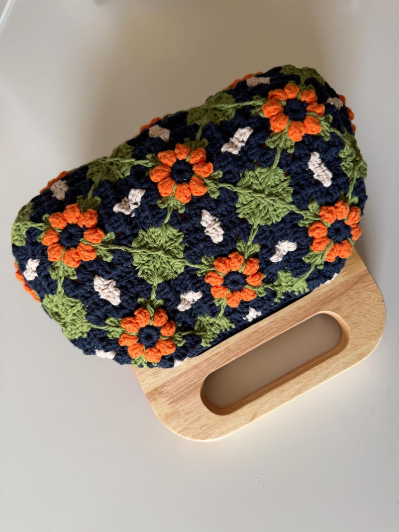 Puede incluir: Un bolso de mano de ganchillo hecho a mano con un estampado floral en azul marino, naranja, verde y blanco. El bolso tiene un asa de madera. El dise&ntilde;o floral incluye flores naranjas, hojas verdes y detalles blancos.
