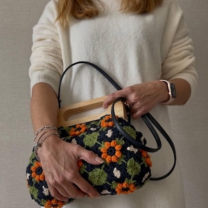 Puede incluir: Bolso de mano de ganchillo con asa de madera y correa de cuero negro. El bolso presenta un fondo azul marino con detalles de flores naranjas y blancas y hojas verdes. Un accesorio elegante.