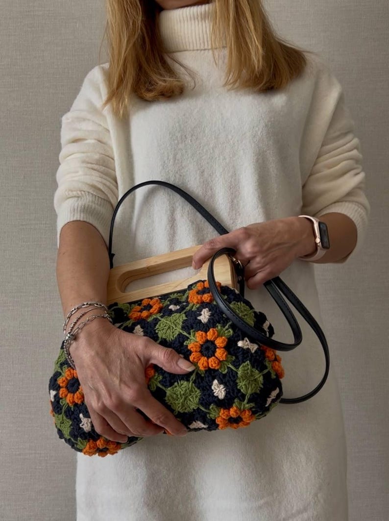 Puede incluir: Bolso de mano de ganchillo con asa de madera y correa de cuero negro. El bolso presenta un fondo azul marino con detalles de flores naranjas y blancas y hojas verdes. Un accesorio elegante.