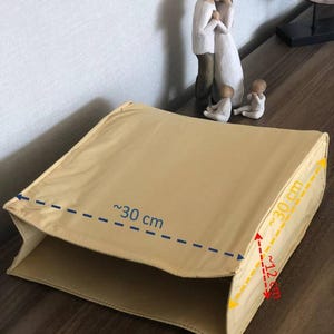 Può includere: Una scatola di stoffa beige di forma rettangolare. La scatola è larga circa 30 cm e alta 12 cm. La scatola è piegata a metà e i bordi sono visibili.