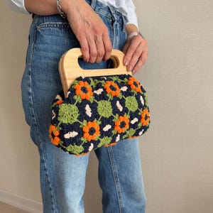 Puede incluir: Un bolso de mano de ganchillo con asa de madera. El bolso presenta un estampado floral en naranja, verde y blanco sobre un fondo azul marino. El asa es de madera clara.