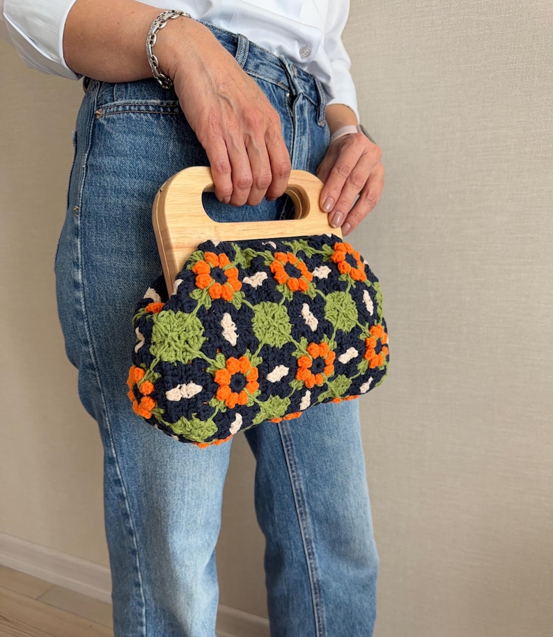 Puede incluir: Un bolso de mano de ganchillo con asa de madera. El bolso presenta un estampado floral en naranja, verde y blanco sobre un fondo azul marino. El asa es de madera clara.