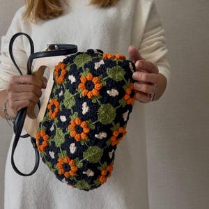 Puede incluir: Un bolso &uacute;nico con asa de madera y correa de cuero negro. El bolso est&aacute; cubierto con un patr&oacute;n de ganchillo de flores naranjas y blancas sobre un fondo azul marino, con hojas verdes.