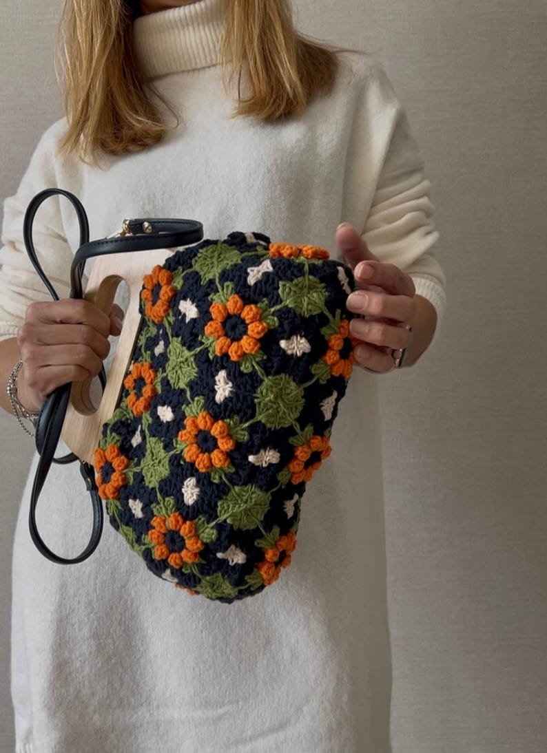 Puede incluir: Un bolso &uacute;nico con asa de madera y correa de cuero negro. El bolso est&aacute; cubierto con un patr&oacute;n de ganchillo de flores naranjas y blancas sobre un fondo azul marino, con hojas verdes.