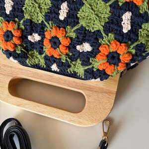 Puede incluir: Bolso de crochet hecho a mano con asa de madera y correa negra. El bolso presenta un estampado floral en azul marino, naranja, verde y blanco. El asa de madera es de color marr&oacute;n claro.
