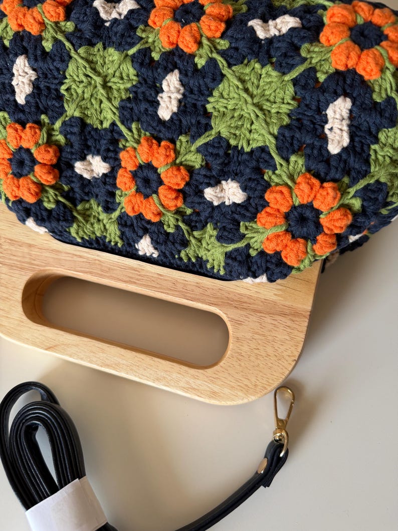 Puede incluir: Bolso de crochet hecho a mano con asa de madera y correa negra. El bolso presenta un estampado floral en azul marino, naranja, verde y blanco. El asa de madera es de color marr&oacute;n claro.