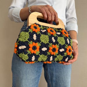 Puede incluir: Un bolso de mano de ganchillo con asa de madera. El bolso presenta un estampado floral en colores azul marino, naranja, verde y crema. El asa es de madera clara. El bolso lo sostiene una persona que lleva una camisa azul claro y vaqueros.