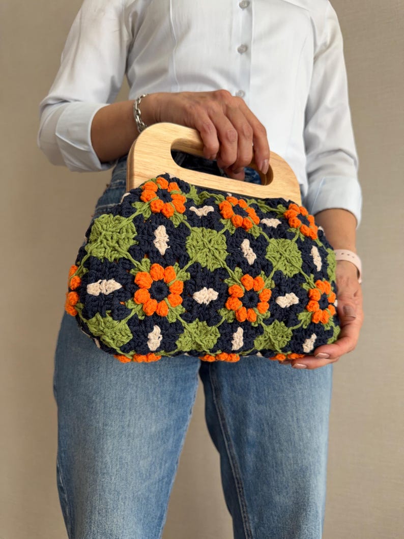 Puede incluir: Un bolso de mano de ganchillo con asa de madera. El bolso presenta un estampado floral en colores azul marino, naranja, verde y crema. El asa es de madera clara. El bolso lo sostiene una persona que lleva una camisa azul claro y vaqueros.