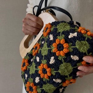 Puede incluir: Bolso de mano de ganchillo con asa de madera y correas de cuero negro. El bolso presenta un estampado floral con detalles en naranja, verde, azul marino y blanco. El bolso lo sostiene una persona que lleva un jersey blanco.