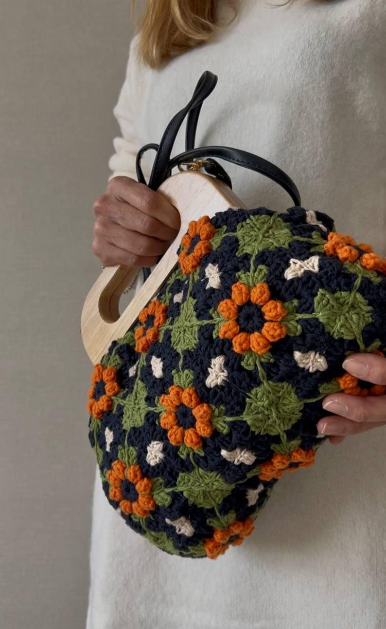 Puede incluir: Bolso de mano de ganchillo con asa de madera y correas de cuero negro. El bolso presenta un estampado floral con detalles en naranja, verde, azul marino y blanco. El bolso lo sostiene una persona que lleva un jersey blanco.