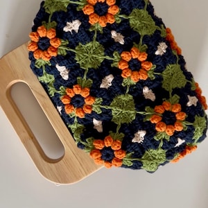 Puede incluir: Bolso de crochet azul marino con asa de madera. El bolso presenta un estampado floral con detalles en naranja, verde y blanco. El dise&ntilde;o de crochet es intrincado y detallado, mostrando una est&eacute;tica hecha a mano.