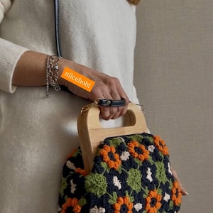 Puede incluir: Bolso de mano de ganchillo hecho a mano con asa de madera. El bolso presenta un estampado floral en naranja, verde y blanco sobre un fondo azul marino. El asa es de madera clara y tiene un agarre de cuero negro.