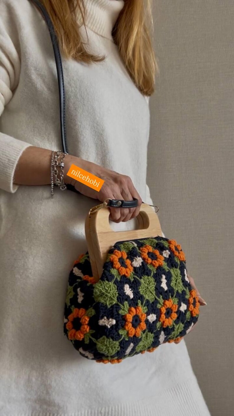 Puede incluir: Bolso de mano de ganchillo hecho a mano con asa de madera. El bolso presenta un estampado floral en naranja, verde y blanco sobre un fondo azul marino. El asa es de madera clara y tiene un agarre de cuero negro.